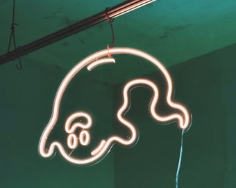 May include: A white neon ghost sign with a smiling face hangs from a ceiling with a red cord. The ghost is outlined in white neon and has a simple, cartoon-like design.