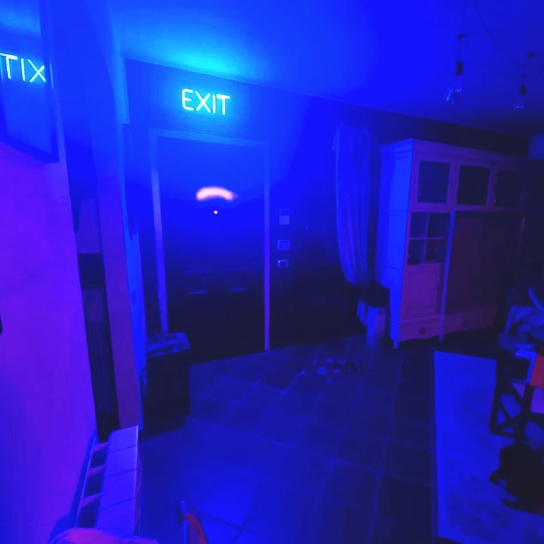 May include: A blue-lit room with a white "EXIT" sign above a door. The room is dimly lit with a wooden cabinet in the background.