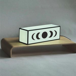May include: A rectangular light box with a pale green surface and a black frame. The front panel features a black graphic design showing the phases of the moon, including four crescent shapes and a central full circle, illuminated from within.