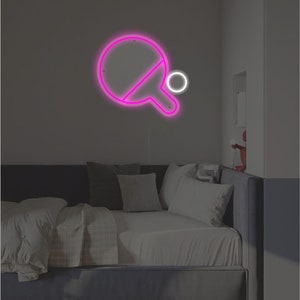 May include: Pink neon sign of a ping pong paddle and ball on a wall above a bed with gray bedding.