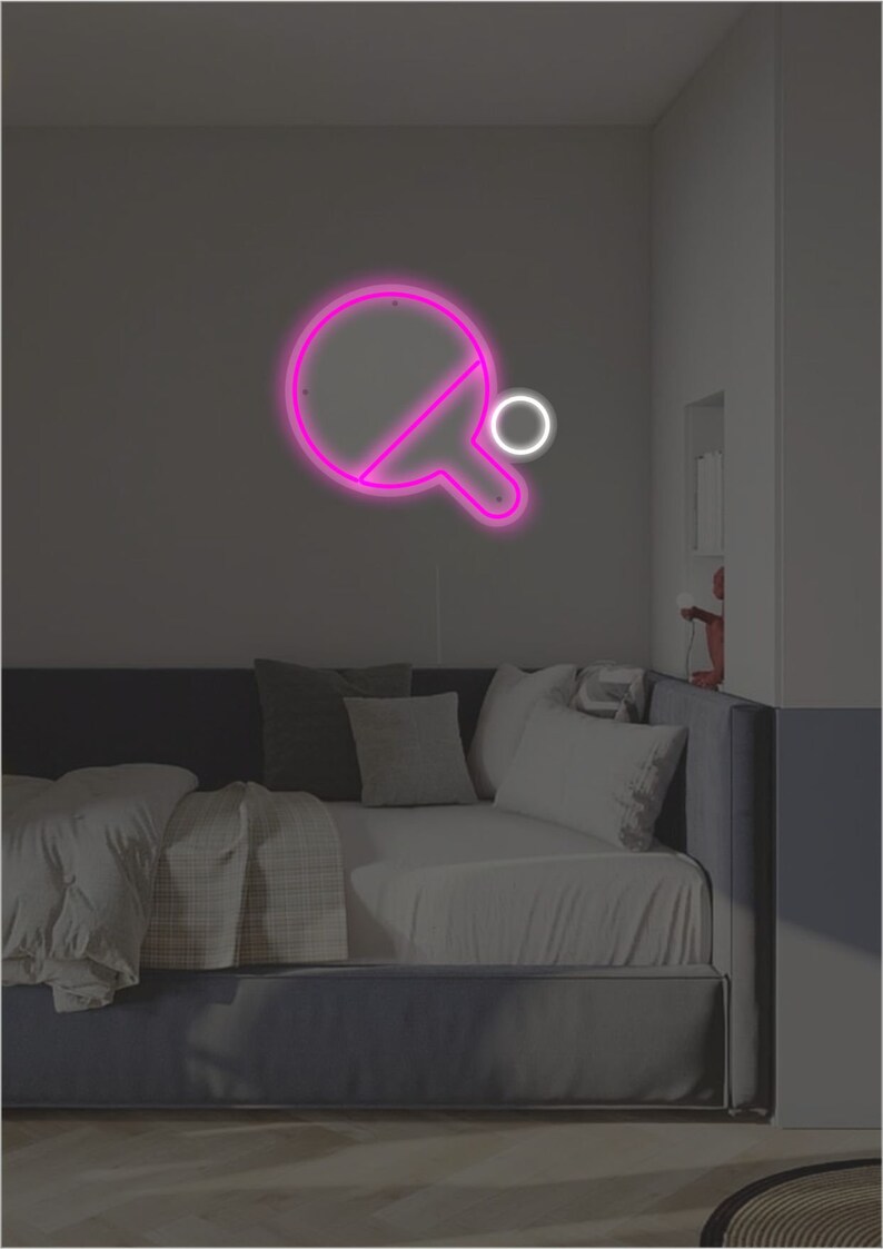 May include: Pink neon sign of a ping pong paddle and ball on a wall above a bed with gray bedding.