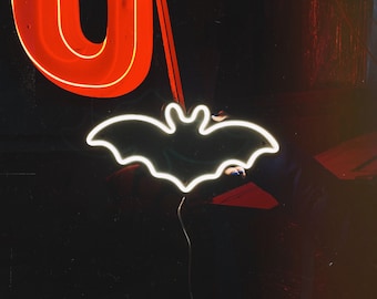 May include: A neon sign shaped like a bat, glowing white against a dark background. A red neon sign shaped like the letter "O" is visible in the upper left corner of the image.