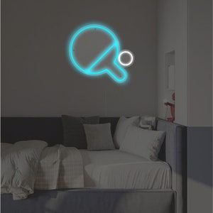 May include: A blue neon sign of a ping pong paddle and ball on a wall above a gray couch with white pillows.