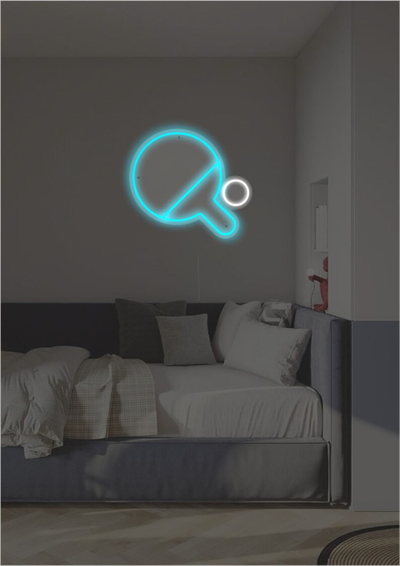 May include: A blue neon sign of a ping pong paddle and ball on a wall above a gray couch with white pillows.