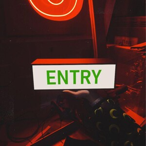 May include: A red neon sign with the word "ENTRY" in green letters on a white background. The sign is lit up and the letters are visible. The sign is in a dark room with a red neon sign in the background.