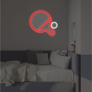 May include: A red neon sign of a ping pong paddle and ball on a wall above a gray couch with white pillows.