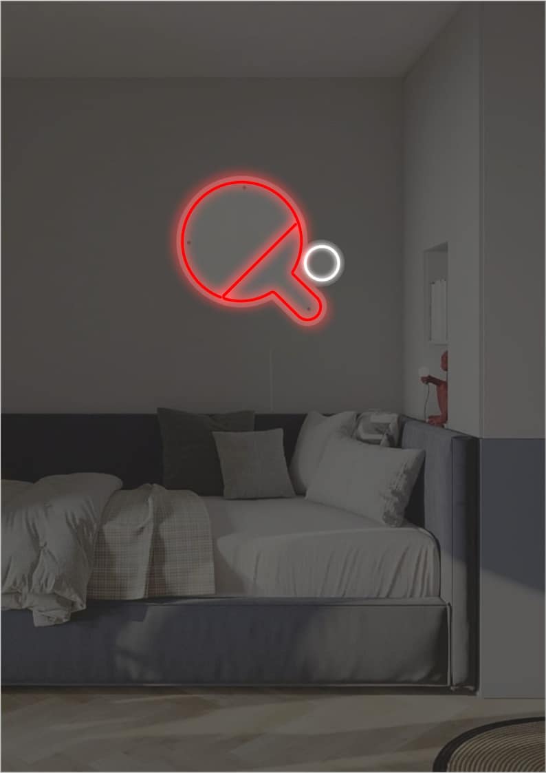 May include: A red neon sign of a ping pong paddle and ball on a wall above a gray couch with white pillows.