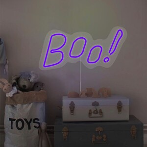 May include: A purple neon "BOO!" sign with an exclamation point stands on a white stand. The sign is placed on top of two decorative trunks. A bag of toys and stuffed animals are in the background. The scene has a Halloween theme.