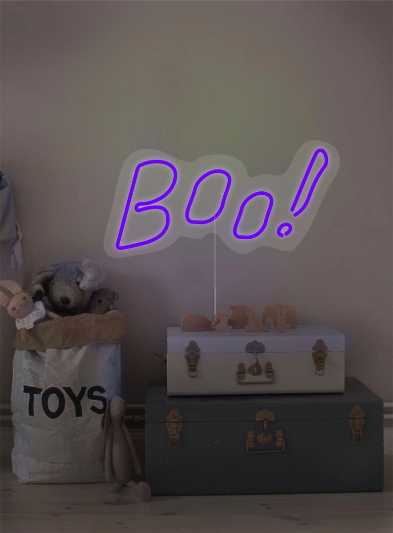 May include: A purple neon "BOO!" sign with an exclamation point stands on a white stand. The sign is placed on top of two decorative trunks. A bag of toys and stuffed animals are in the background. The scene has a Halloween theme.