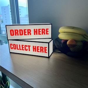Op de afbeelding: Twee rechthoekige lichtbakken met zwarte frames en witte vlakken. De bovenste doos leest "ORDER HERE" in rode letters. De onderste doos leest "COLLECT HERE" in rode letters. De dozen staan op een houten oppervlak, met een fruitmand op de achtergrond.