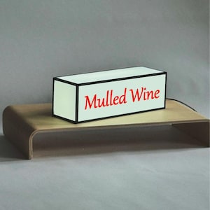 May include: A rectangular white sign with the words "Mulled Wine" in red lettering, framed by a black border. The sign sits on a curved wooden stand, with a gray background.