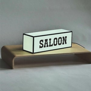 May include: A rectangular light box with the word "SALOON" in bold black letters. The box has a white face and black trim, sitting on a curved wooden stand. The background is a solid gray.