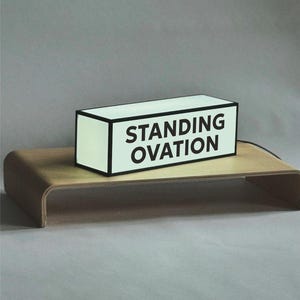 以下が含まれることがあります： 「STANDING OVATION」の文字が黒い太字で書かれた白い長方形のライトボックス。ライトボックスは黒い縁取りがあり、曲がった木製のスタンドの上に置かれています。背景は無地のグレーです。