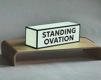 Puede incluir: Una caja de luz rectangular con superficie verde pálido y bordes negros, que presenta el texto STANDING OVATION en letras mayúsculas negras en la parte frontal. La caja está iluminada y se apoya sobre un soporte de madera curvado.