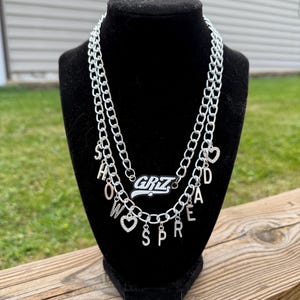 以下が含まれることがあります： 2層のシルバーチェーンネックレス。上層には白黒の「GRIZ」という文字が入った長方形のペンダントが付いています。下層には「SHOW HOW SPREAD」の文字が個別の文字チャームとハートチャームで綴られています。