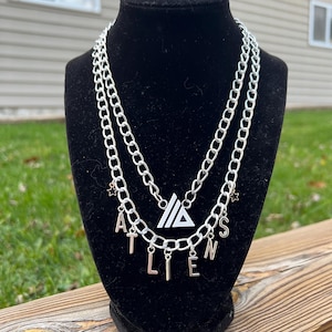 May include: Silver chain necklace with a triangle pendant and the word "ALIENS" spelled out with charms. The necklace is on a black display stand.