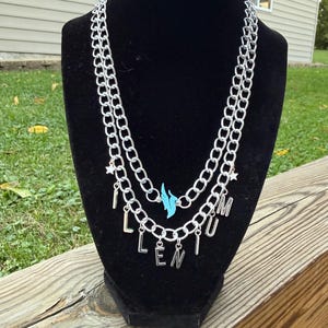 May include: A silver chain necklace with three layers, displayed on a black mannequin. The necklace has the letters "LENIUM" hanging from the bottom chain, a blue bird charm, and star charms.