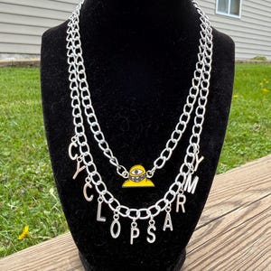 Op de afbeelding: Een zilveren ketting met het woord "CYCLOPS ARMY" in afzonderlijke letters. De ketting heeft een gele hanger met een cartoon cyclops ontwerp.