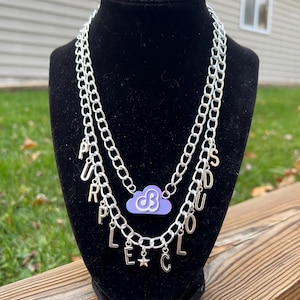 May include: A silver chain necklace with multiple charms, including the letters "PURPLE CLOUDS" and a purple cloud charm with a white "B" inside.