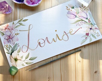 May include: A watercolour painting of the name "Louise" in gold script with pink and white flowers and green leaves around the name.