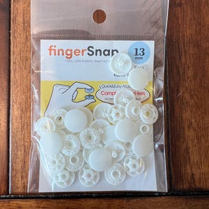 May include: A clear plastic bag containing white fingerSnap fasteners. The package includes multiple snap fasteners, with the text "fingerSnap" and "13 mm" visible. The fasteners are tool-less plastic snap fasteners.