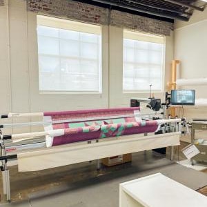 May include: A longarm quilting machine with a quilt in progress. The quilt features a geometric design in shades of purple, green, and white. The machine is in a well-lit workshop with large windows and a computer screen.