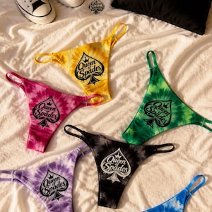 May include: A collection of tie-dye thongs in various colors, including yellow, pink, green, black, purple, and blue, each featuring a "Queen of Spades" graphic. The image also shows a black pillow, a silver necklace, and white sneakers.