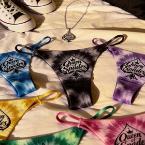 May include: A collection of tie-dye thong underwear in various colors, including blue, black, yellow, green, and pink, each with the "Queen of Spades" logo. A matching necklace and a black pillow with the same logo are also visible.