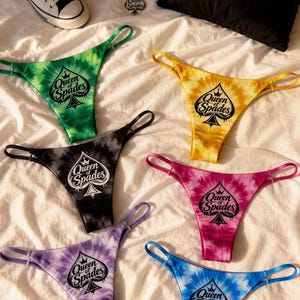 May include: A collection of tie-dye thong underwear in various colors, including green, yellow, black, pink, purple, and blue, each with a spade logo and the text "Queen of Spades". Also, a black pillow, a necklace, and white sneakers.