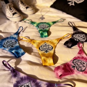 May include: A collection of tie-dye thong underwear in various colors, including blue, yellow, green, black, pink, and purple, each with the text "Queen of Spades" and a spade symbol. A black pillow with a spade symbol and white sneakers are also visible.