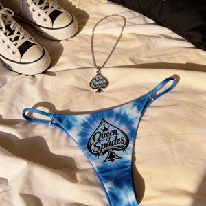 May include: A blue tie-dye thong with the text "Queen of Spades" and a spade graphic, a silver necklace with a matching pendant, a black pillow with the same design, and white high-top sneakers.