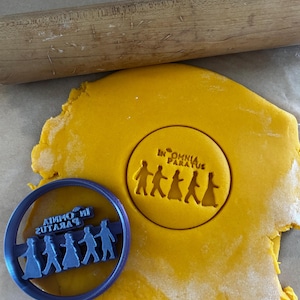 May include: A blue cookie cutter with a circular design and the text "IN OMNIA PARATUS" and a group of people. A yellow dough with the same design is next to the cutter. A wooden rolling pin is in the background.