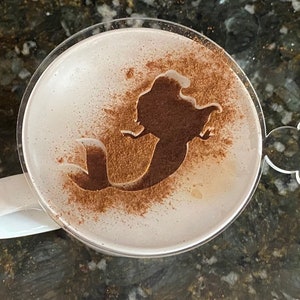 May include: A white mug with a clear lid, the lid has a Mickey Mouse ear design. The mug has a light brown design of a mermaid on the surface of the drink inside.