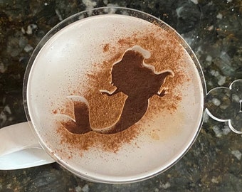 May include: A white mug with a clear lid, the lid has a Mickey Mouse ear design. The mug has a light brown design of a mermaid on the surface of the drink inside.