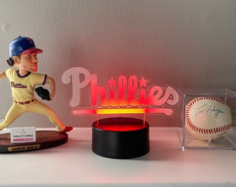May include: A red and white illuminated sign with the word "Phillies" in a stylized font. The sign is on a black base and is lit from below. There is a baseball figurine and a baseball in a display case to the left and right of the sign.