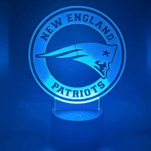 May include: A blue-lit New England Patriots logo, featuring a circular design with the team name and a stylized head and star emblem. The logo is mounted on a clear base, creating a glowing effect against a blue background.