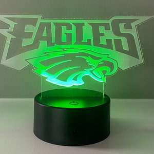 May include: A green LED night light with a clear acrylic base featuring the Philadelphia Eagles logo. The logo is a stylized eagle head with the word "EAGLES" above it.