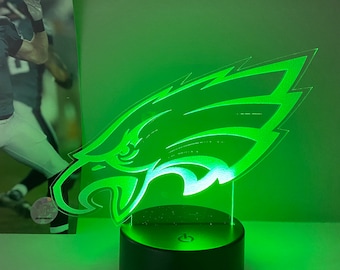 Pode incluir: Uma lâmpada de acrílico verde brilhante com o logotipo dos Philadelphia Eagles. O logotipo é iluminado por luzes LED verdes e fica em uma base preta. Uma foto de um jogador de futebol americano está ao fundo.