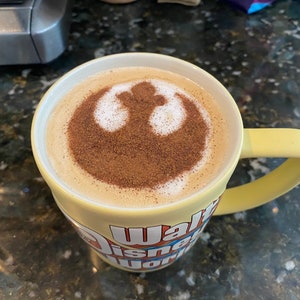 May include: A yellow ceramic mug with a white "Walt Disney World" logo and a cinnamon-dusted latte featuring a white Rebel Alliance symbol.