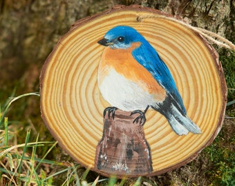 May include: A hand-painted wooden ornament depicting a bluebird perched on a branch. The bird is painted in shades of blue, orange, and white, against a natural wood grain background.