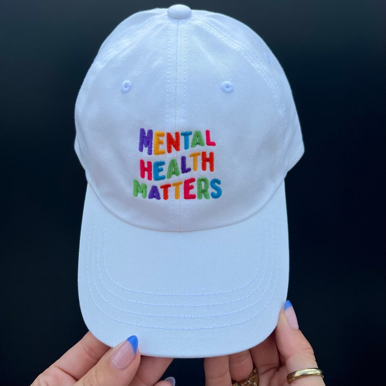 Hands holding a white dad hat with Mental Health Matters embroidered in colorful letters.