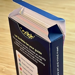 May include: A partially open box of "Scribb" cards. The box is dark blue with white text that reads "Let Scribble be your guide" and "where art and mind meet." The cards are visible inside the box.