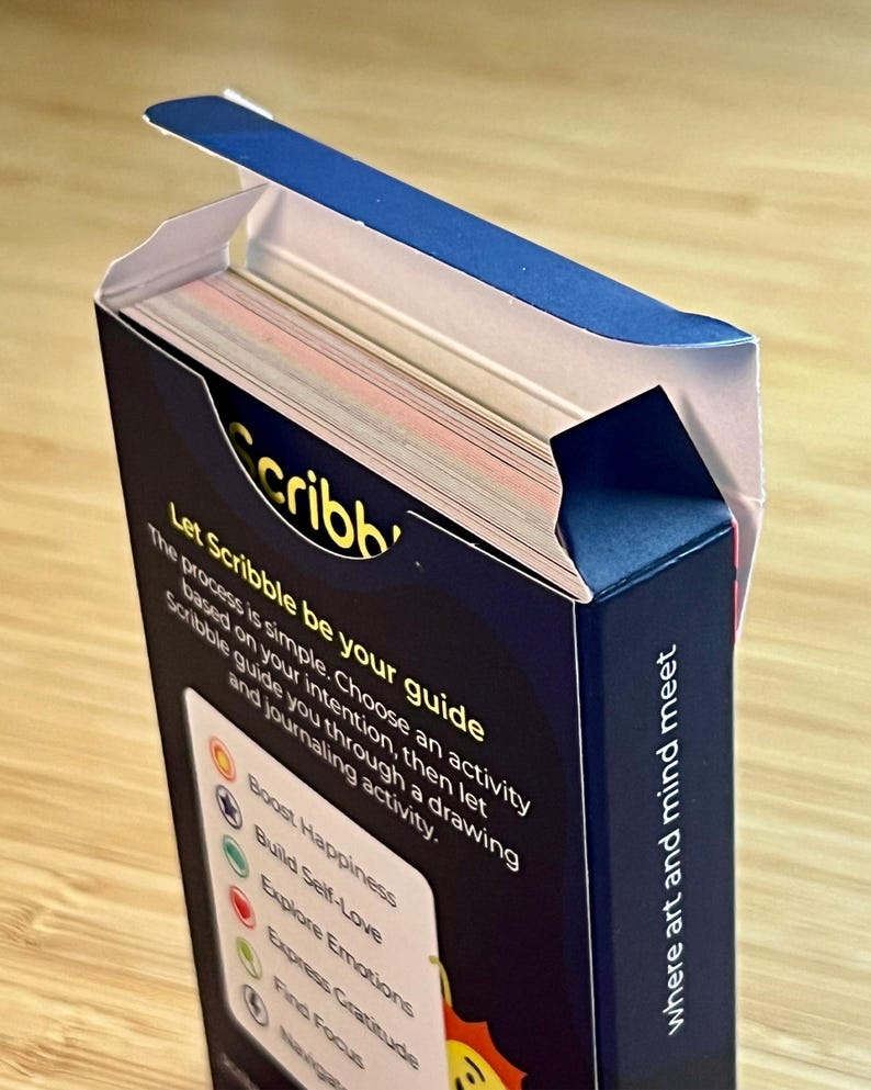 May include: A partially open box of "Scribb" cards. The box is dark blue with white text that reads "Let Scribble be your guide" and "where art and mind meet." The cards are visible inside the box.
