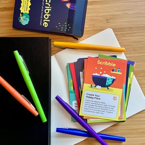 May include: An overhead shot of a collection of stationery items. The image features a black notebook, a deck of cards labeled "Scribble", and several colorful pens in orange, green, yellow, purple, and blue. The cards have colorful illustrations and text.