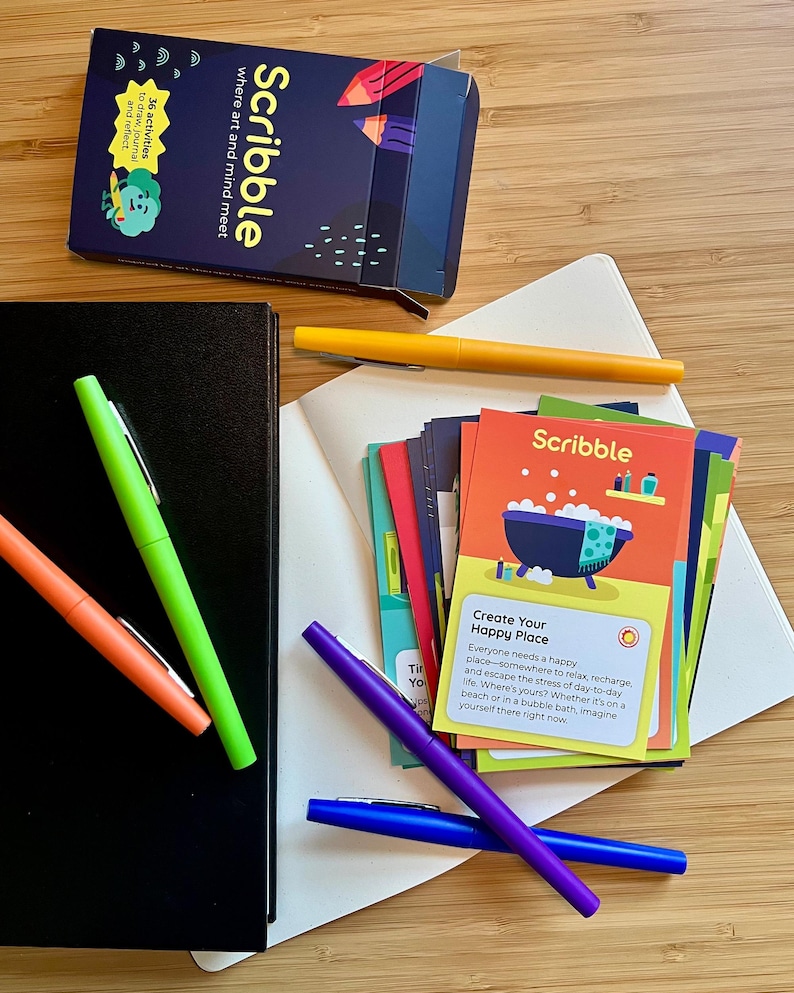 May include: An overhead shot of a collection of stationery items. The image features a black notebook, a deck of cards labeled "Scribble", and several colorful pens in orange, green, yellow, purple, and blue. The cards have colorful illustrations and text.