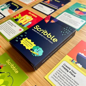 May include: A navy blue box of "Scribble" cards with the text "where art and mind meet" and "36 activities to draw, journal and reflect." Several colorful cards are scattered around the box, featuring illustrations and text.