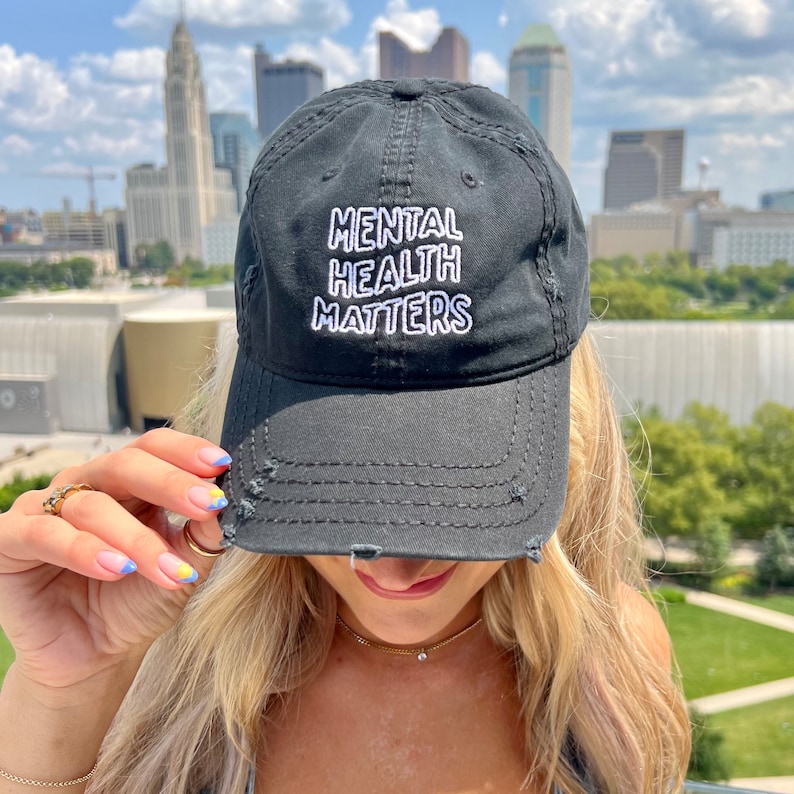 A model wearing a black distressed dad hat that is embroidered with the text Mental Health Matters. She is standing in front of the Columbus, OH city skyline.