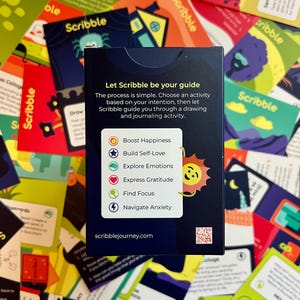 May include: A deck of "Scribble" cards with a navy blue box featuring text and icons for activities like boosting happiness and exploring emotions. The cards are scattered around the box, displaying colorful illustrations.