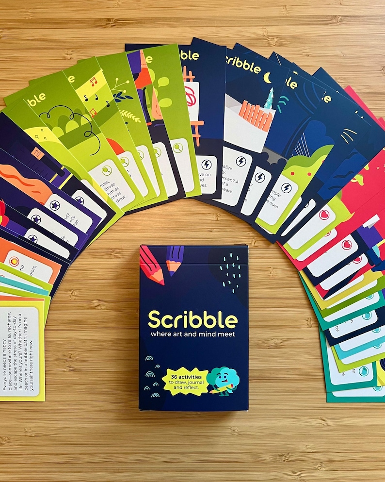 May include: A deck of "Scribble" cards with a navy blue box, featuring the text "Scribble where art and mind meet". The box also states "36 activities to draw, journal and reflect". The cards are arranged in an arc, with colorful illustrations.