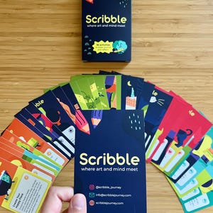 May include: A deck of "Scribble" cards with the tagline "where art and mind meet." The navy blue box and cards feature colorful illustrations and text. The deck includes 36 activities to draw, journal, and reflect. The card in the foreground displays social media information.